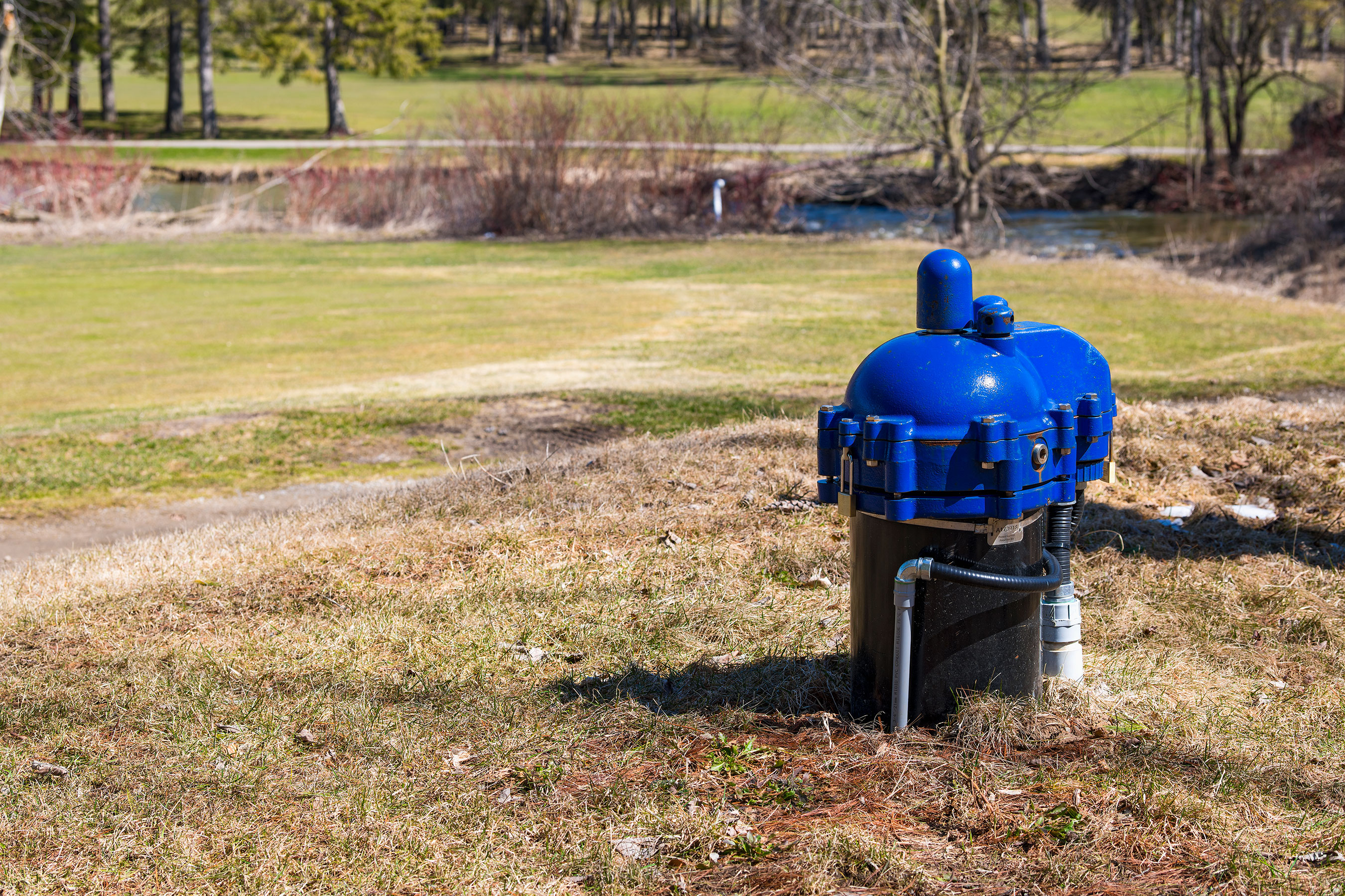 A blue well head located in a grassy field with trees and a small body of water in the background on a sunny day.